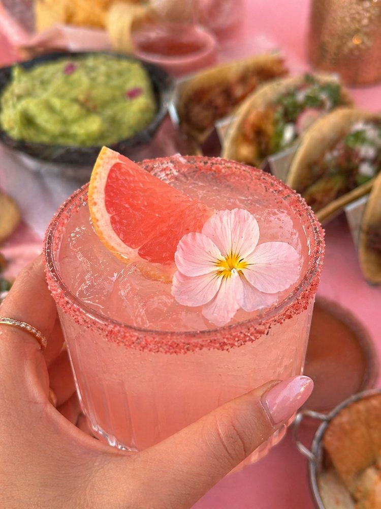 A pink salt-rimmed margarita held up over tacos and guacamole