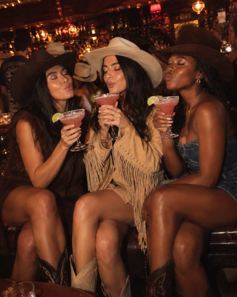 Three friends in cowboy hats sitting on a bench in a bar, each holding a frozen margarita