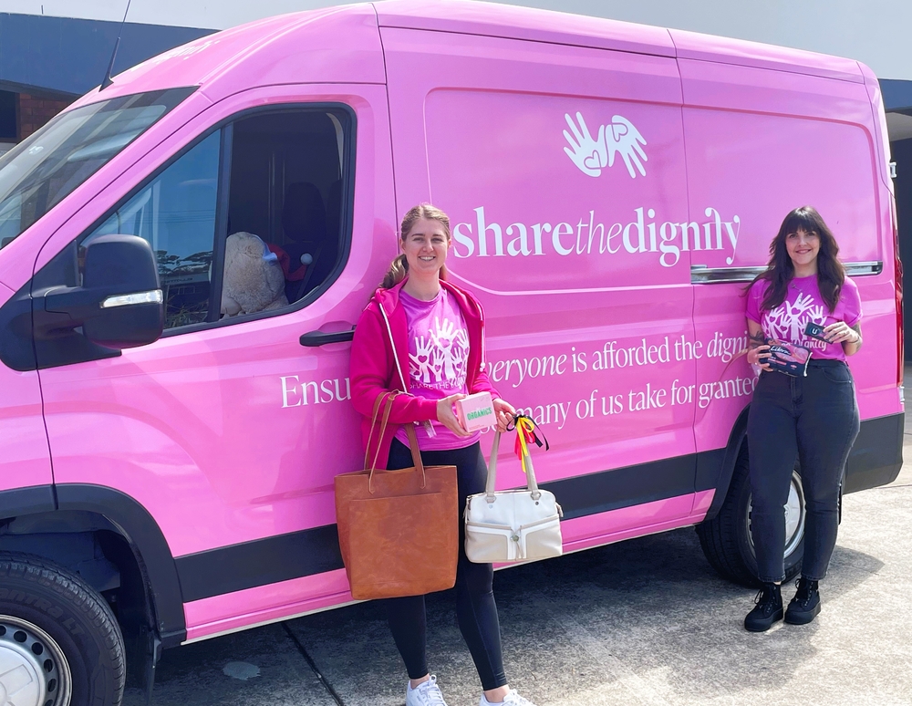 Two women standing beside the Share the Dignity delivery van, each holding a handbag packed with essentials.