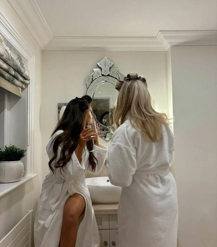 Two friends in robes and hair rollers getting ready together at the bathroom mirror