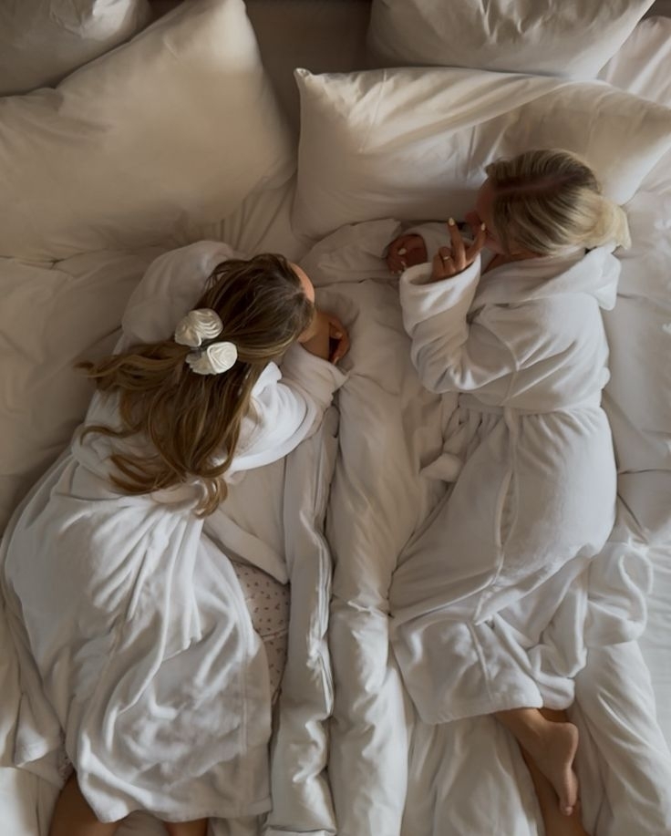Two friends in robes sprawled across fluffy white bedding the morning after a sleepover
