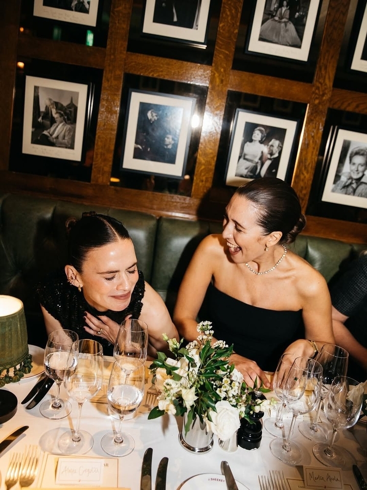 Two friends laughing over dinner at a white tablecloth table with flowers