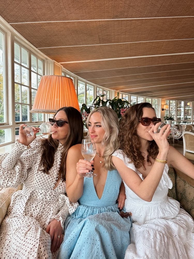 Three friends in summer dresses sipping wine in a bright, airy dining room