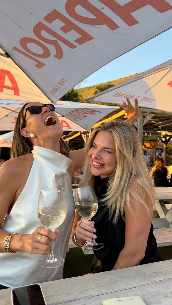 Two friends laughing with glasses of white wine at a sunny outdoor bar