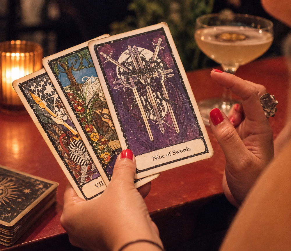 Tarot cards fanned out in one hand beside a cocktail and candlelight