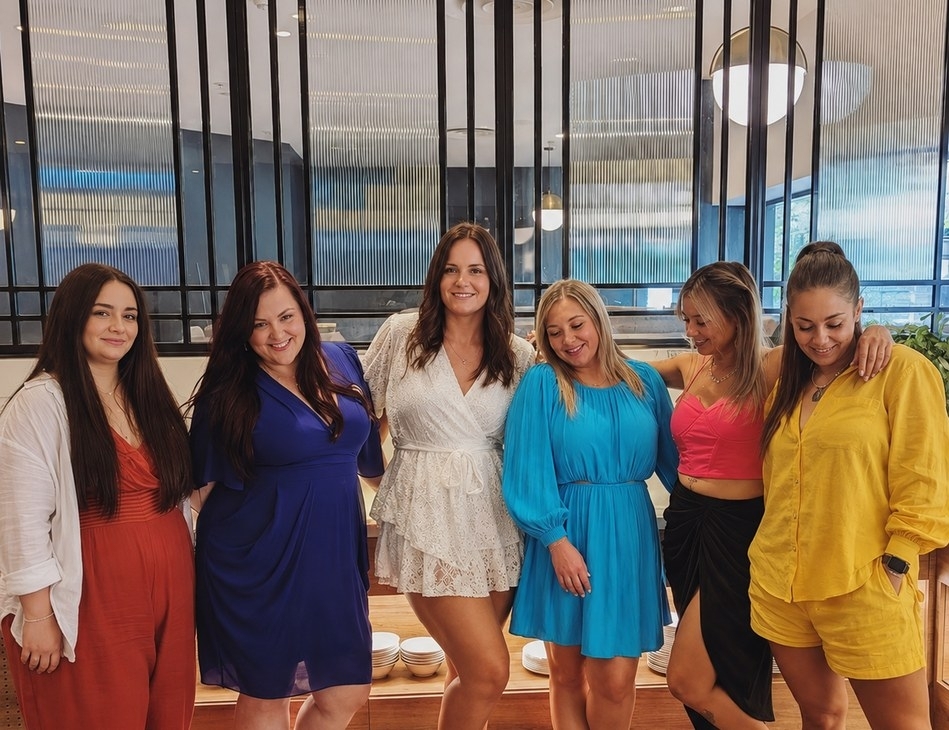 Six friends in colourful outfits with their arms around each other in a bright restaurant
