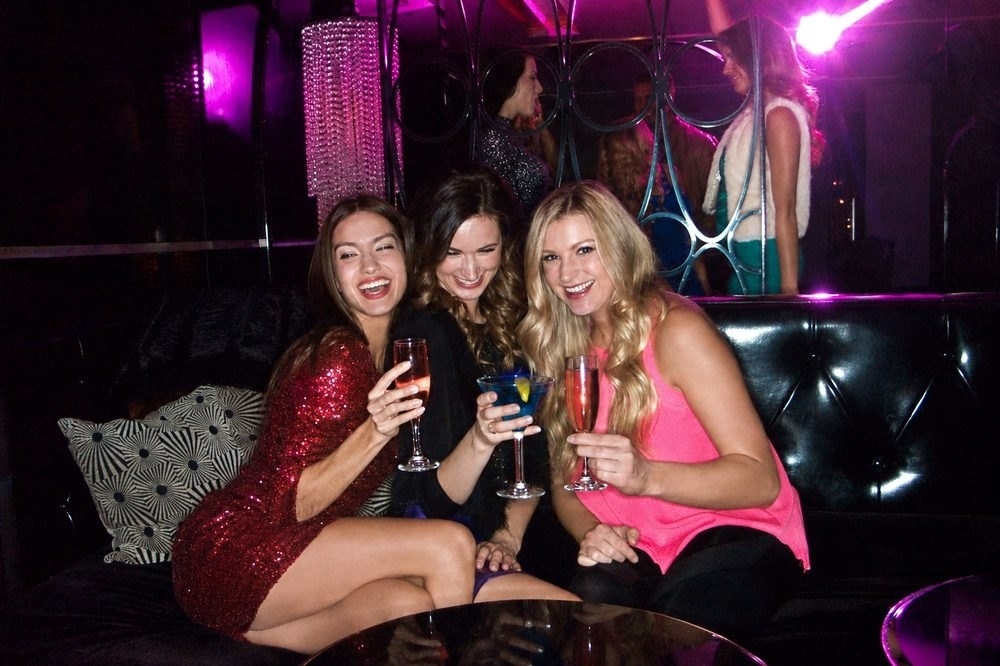 Three friends laughing and raising drinks in a nightclub booth