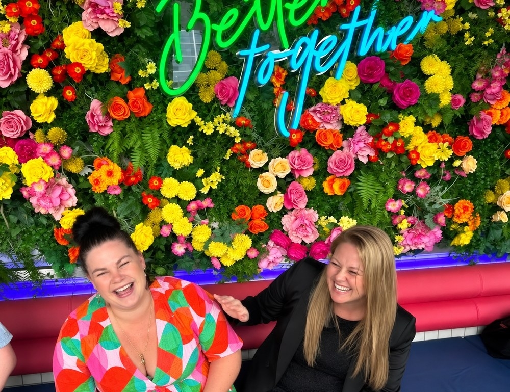Two friends laughing in a pink booth under a flower wall