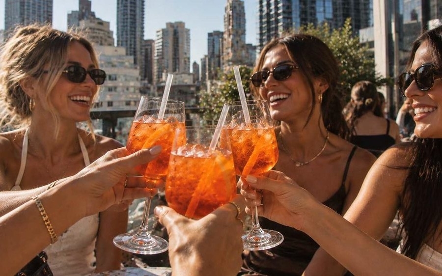 Friends clinking orange spritzes on a sunny rooftop with city buildings behind