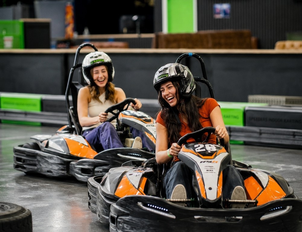 Two friends in helmets laughing as they race e-karts around an indoor track