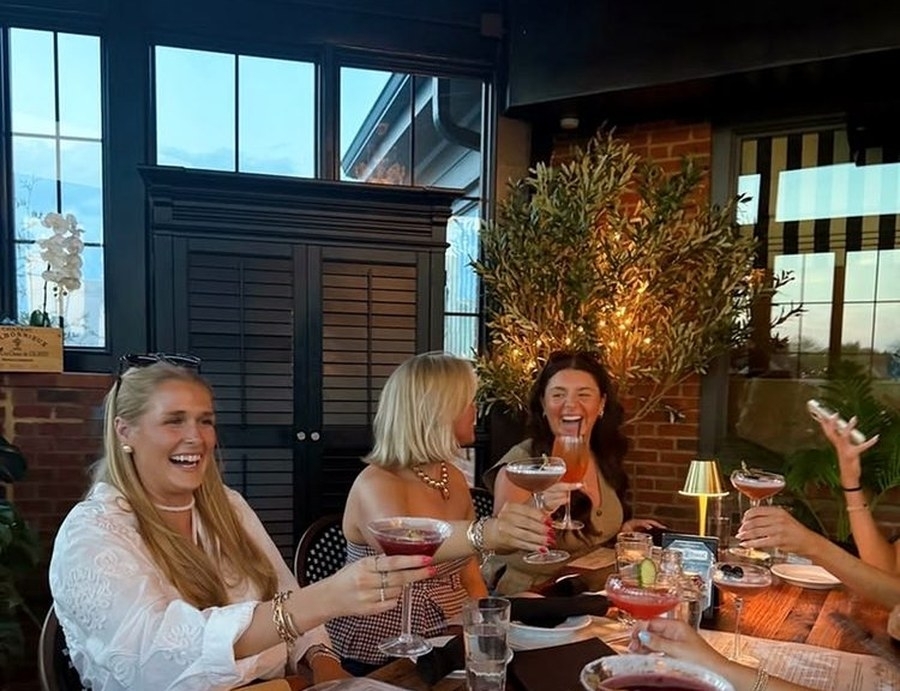 Friends laughing over cocktails at a terrace table with warm lamps and dark green shutters behind