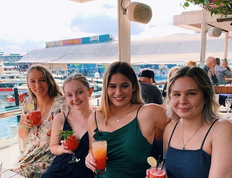 Four friends holding colourful cocktails at a sunny table on the water, with a wharf and boats behind them