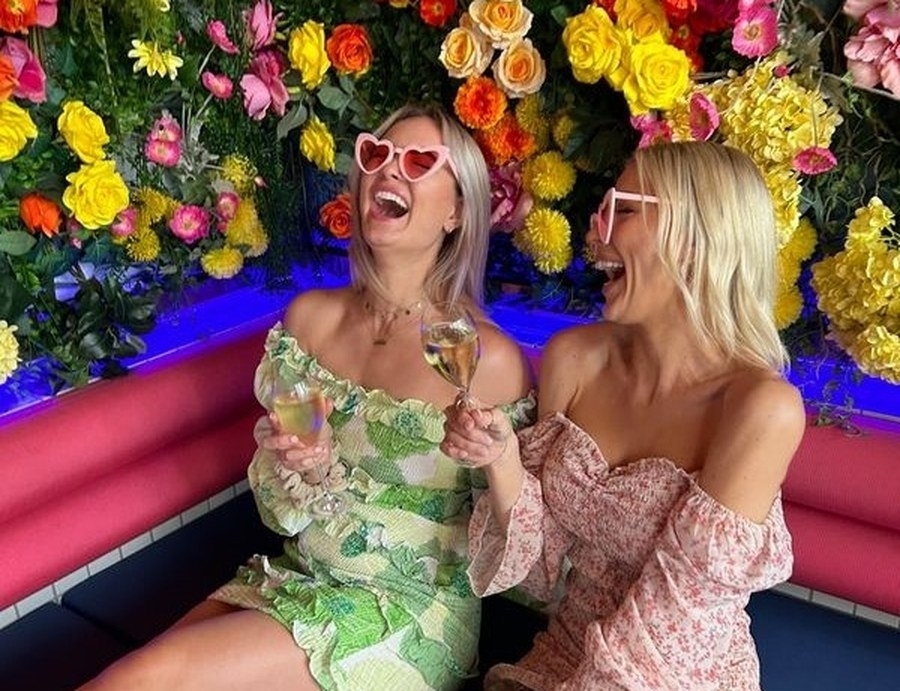 Two friends in heart sunglasses laughing in a flower wall booth