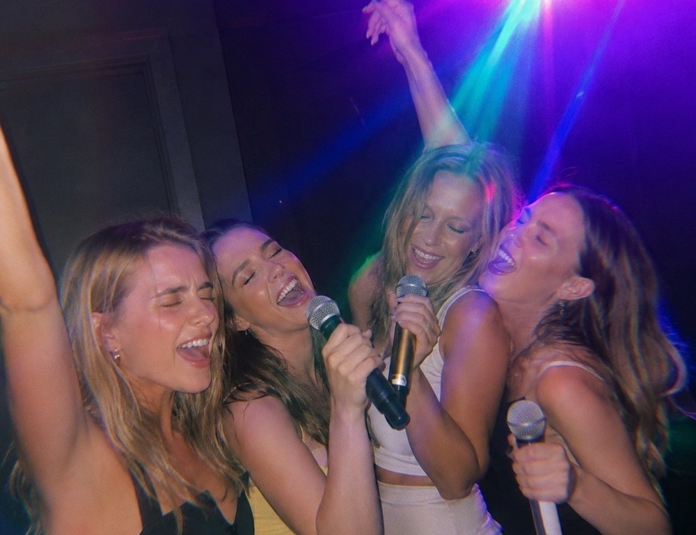 Four friends singing into microphones with their eyes shut under party lights