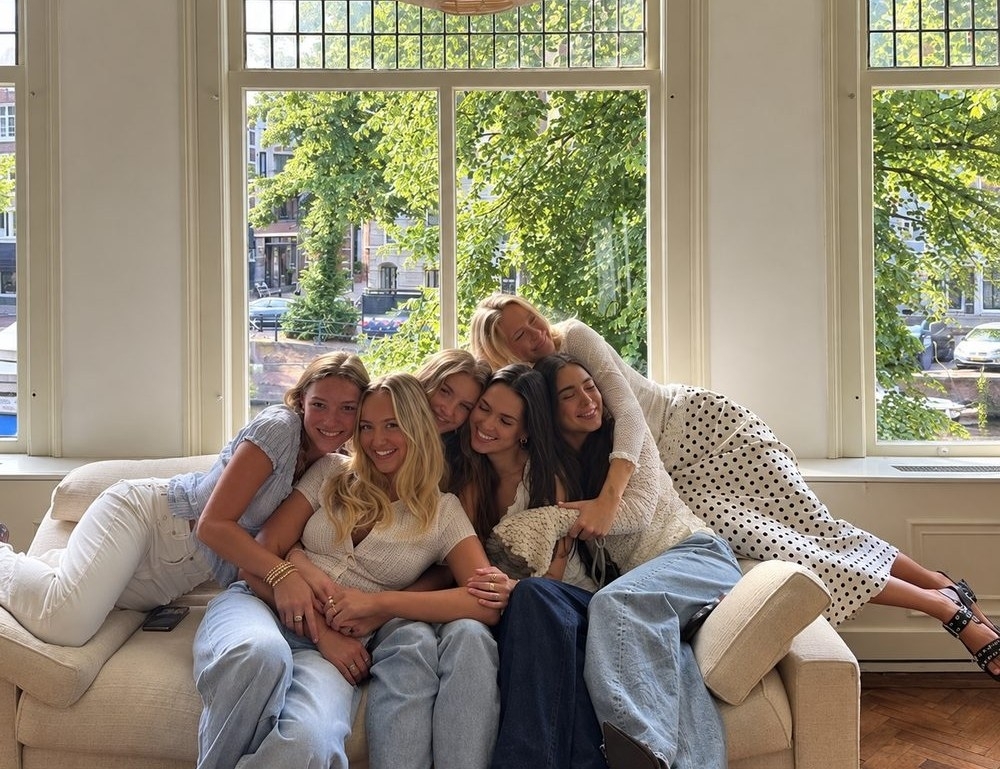 Five friends squeezed together and laughing on a cream sofa in front of a tall sunny window