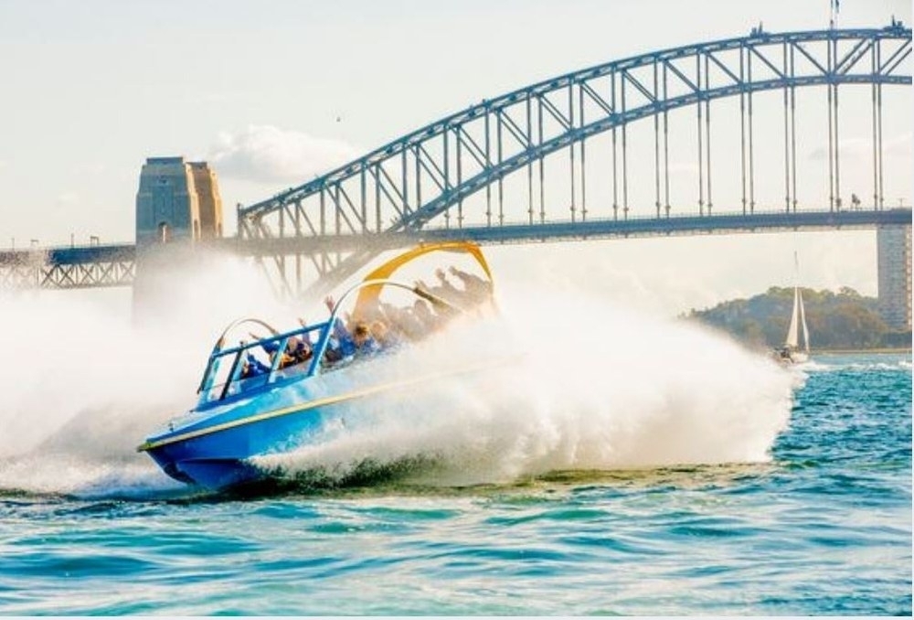 A jet boat throwing up spray on the blue water in front of a big steel arch bridge