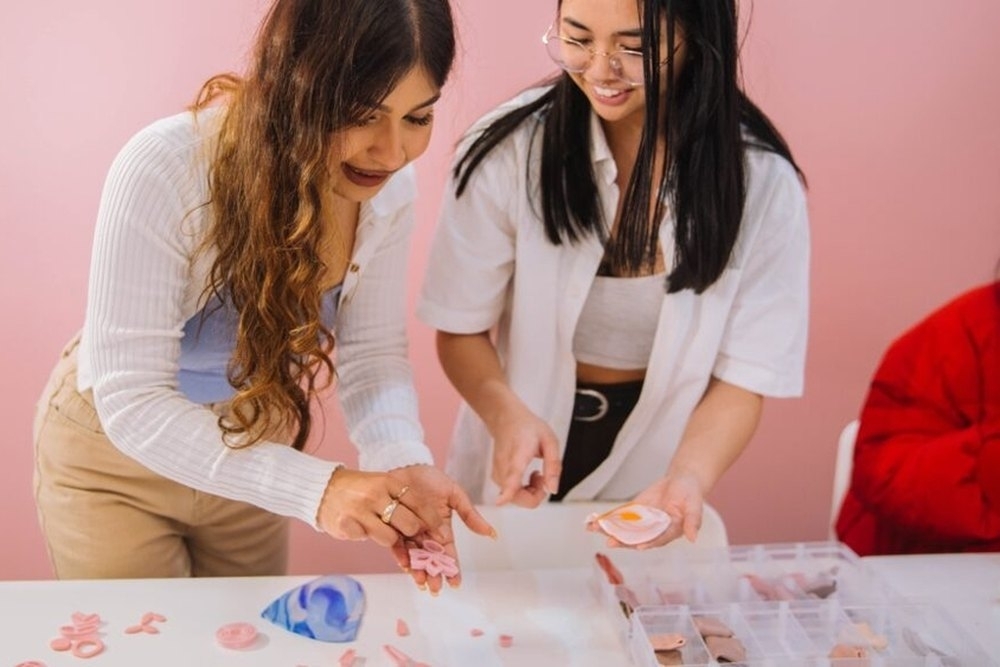 Two friends leaning over the table shaping clay in a pink studio