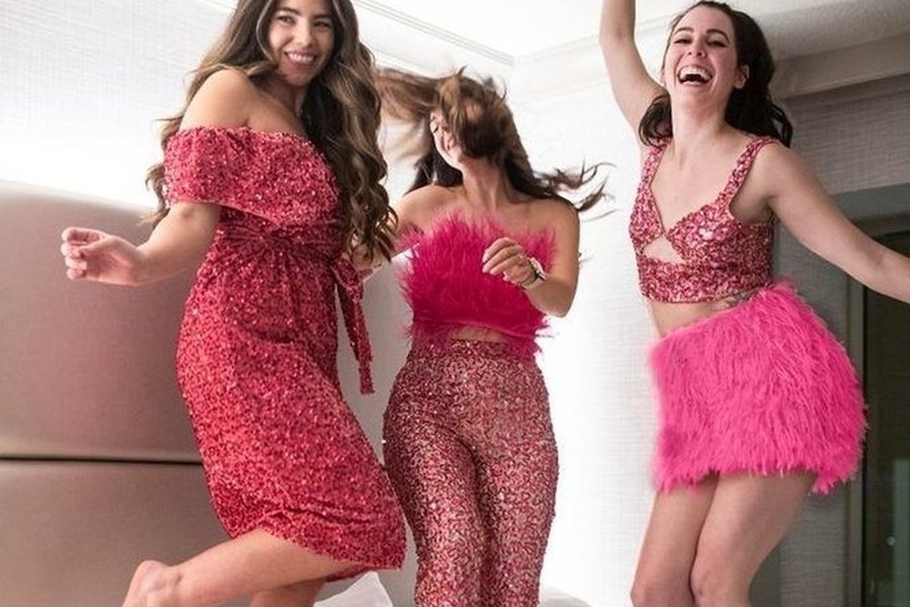 Three friends in pink sequins and feathers jumping on a bed, laughing