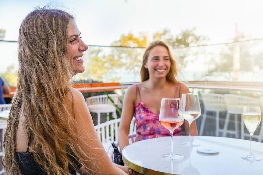Two friends laughing over glasses of wine at a sunny rooftop table