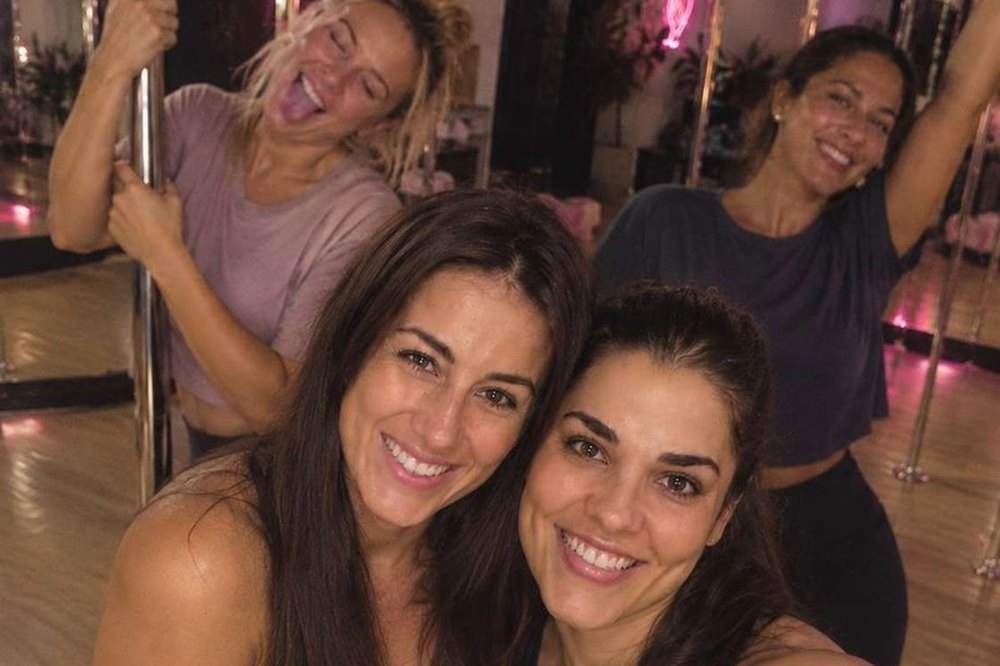 Friends grinning in a group selfie by the poles after their pole dancing class