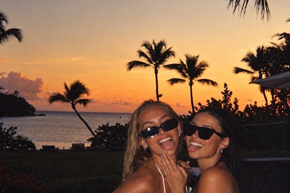 Two friends in sunglasses laughing together under palm trees at sunset
