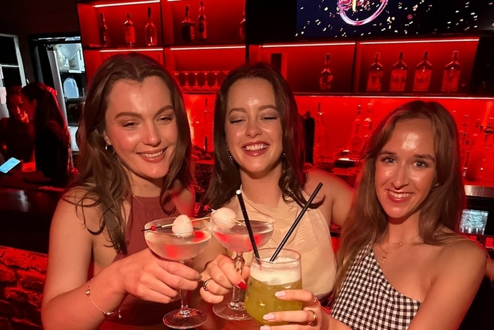 Three friends raising their drinks at a red-lit bar lined with bottles