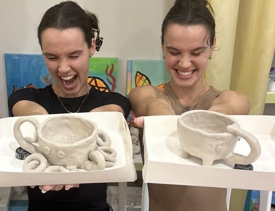 Two friends laughing as they hold up the clay mugs they made