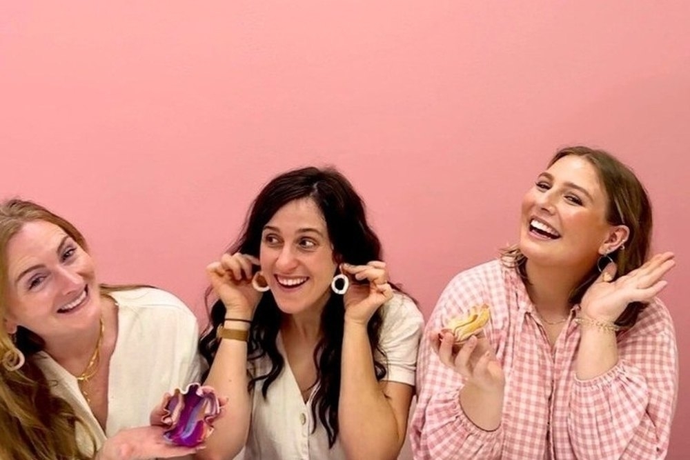 Three friends laughing and showing off their clay pieces against a pink wall