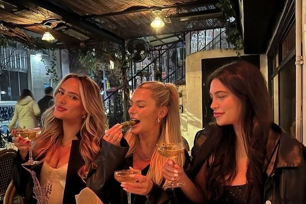 Three friends laughing with drinks under the lights at a leafy bar at night