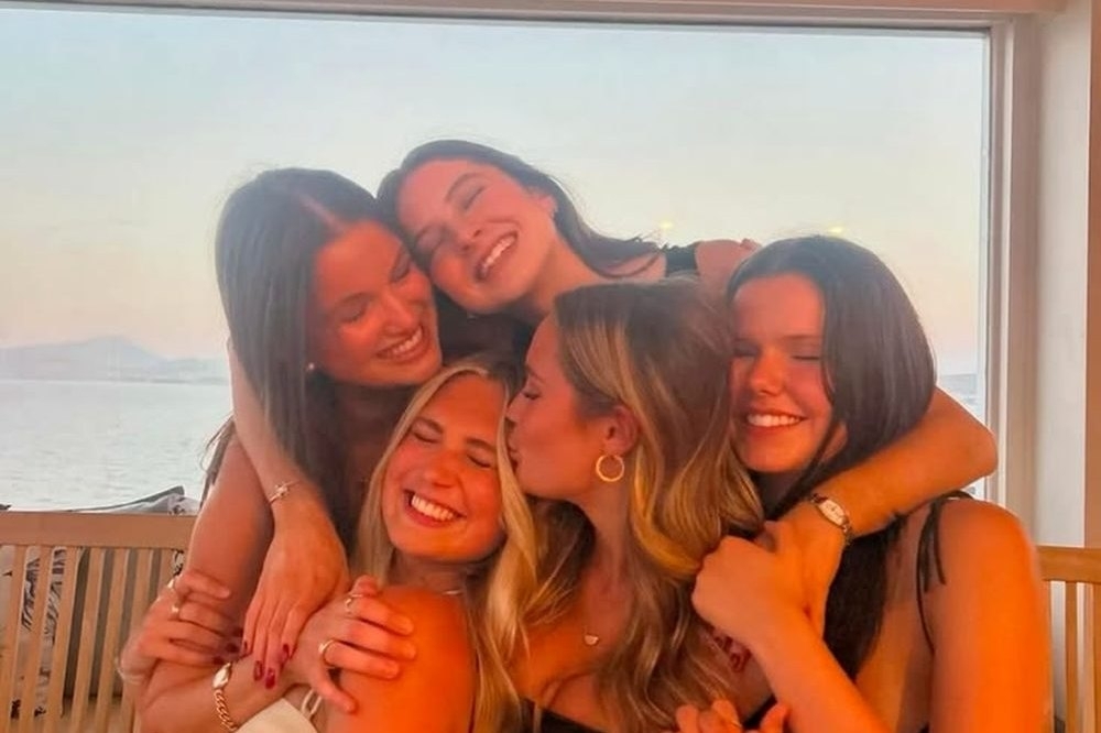 Five friends in a tight group hug by the window at golden hour