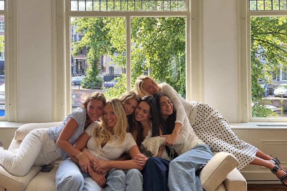 Five friends squeezed together and laughing on a cream sofa in front of a big bright window