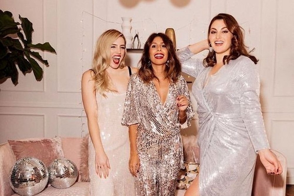 Three friends in sequins laughing together in front of a pink lounge and disco balls