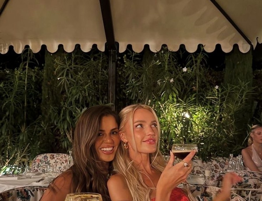 Two friends with drinks under a scalloped umbrella at a leafy garden bar at night