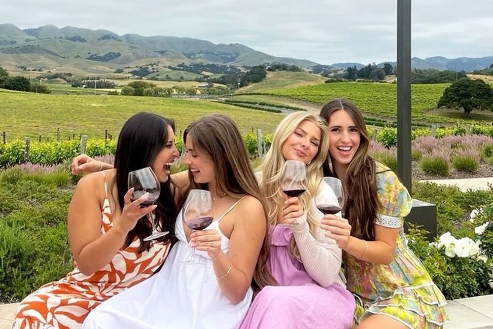 Four friends laughing with red wine on a terrace wall overlooking the vines