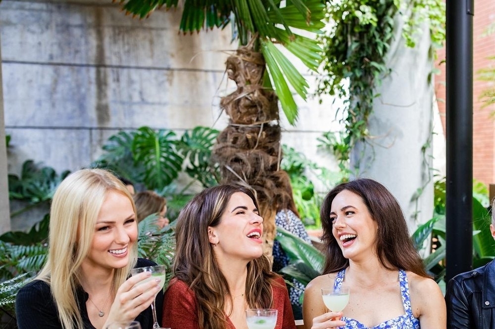 Three friends laughing over lunch and wine at a long leafy table