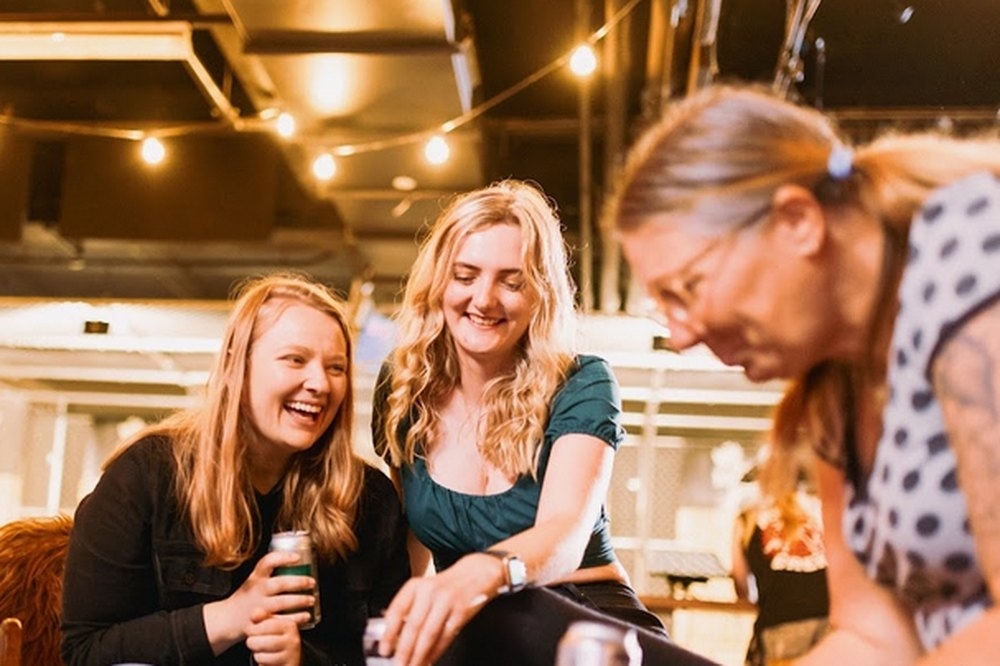 Three friends laughing over a share plate and drinks under string lights