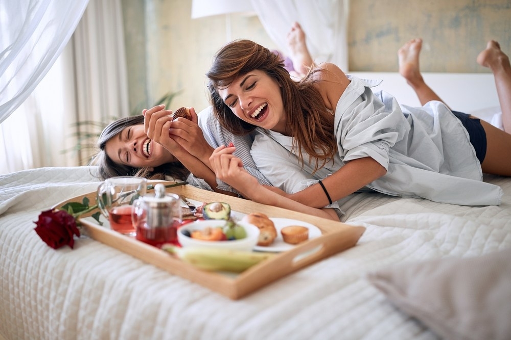 Two friends laughing on the bed beside a breakfast tray the morning after