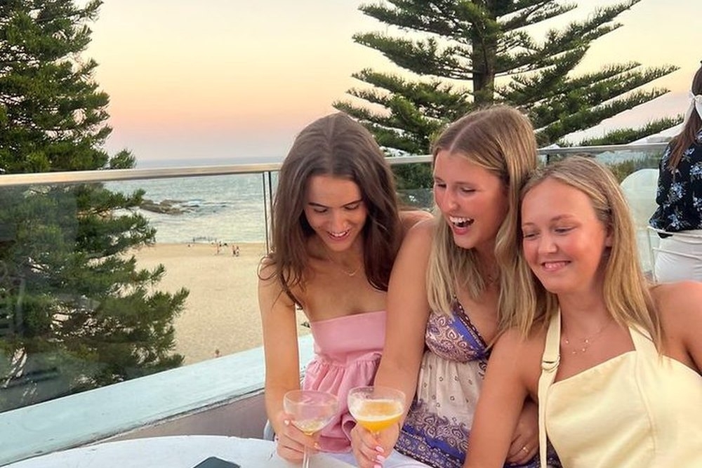 Three friends laughing with drinks on a balcony above the beach at sunset