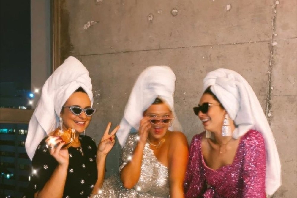 Three friends in towel turbans and sequin dresses snacking mid glam