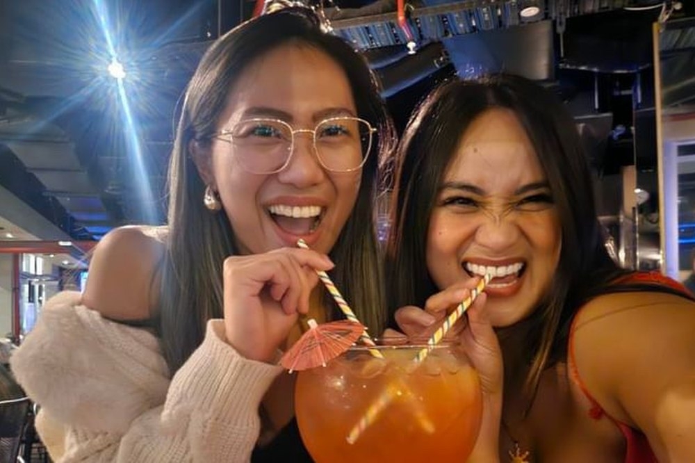 Two friends laughing as they share a big cocktail through two straws at a lively bar