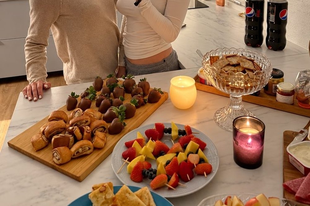 Two friends chatting beside a candlelit grazing spread in the kitchen