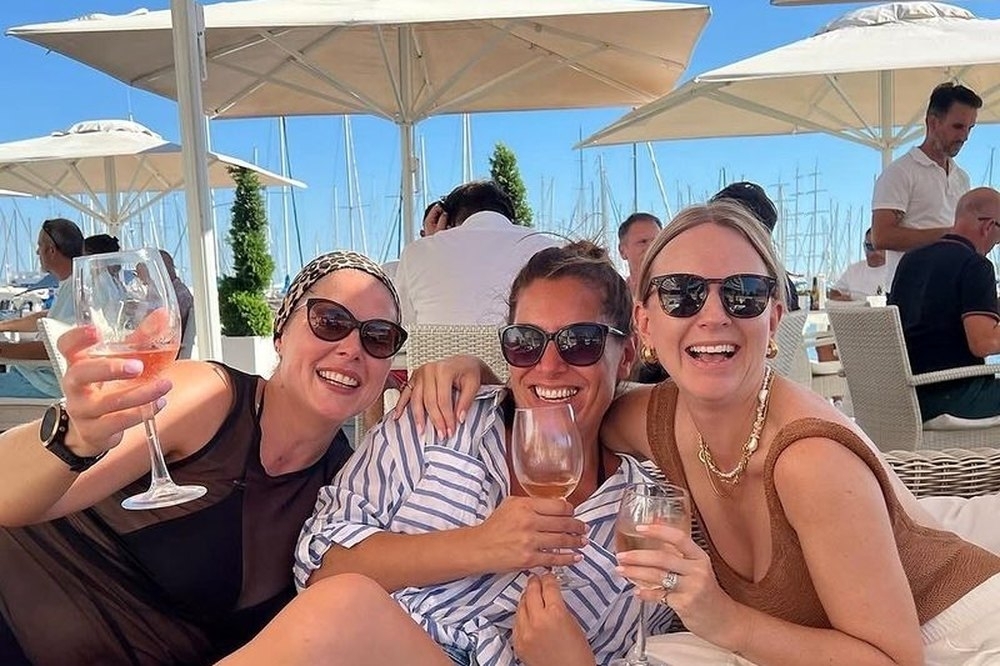 Three friends in sunglasses laughing with drinks under the umbrellas at a beach club