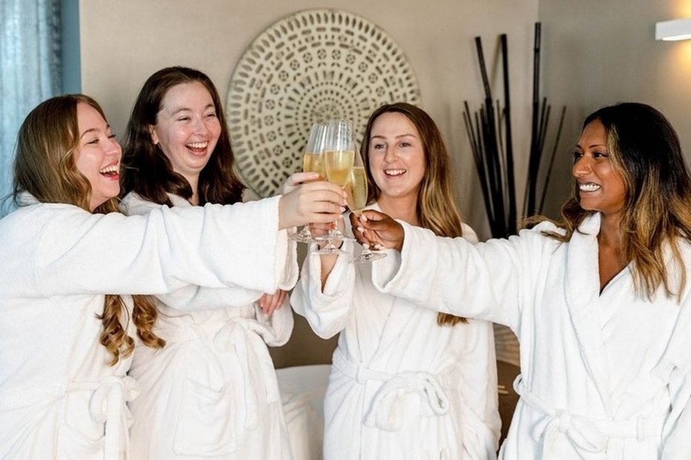Four friends in white robes laughing and toasting with bubbly at a day spa